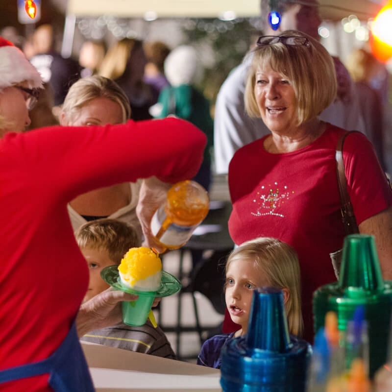 A Tropical Sno independent dealer is pouring peach-flavored syrup over fluffy shaved ice from a Swan Shaved Ice machine, and a queue of customers is waiting. A mother smiles broadly while her daughter looks on in eager anticipation of tasting the frozen treat.