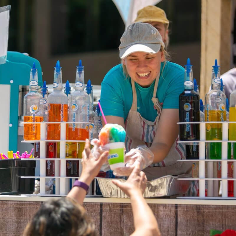 A Tropical Sno dealer smiles broadly as she hands out a cherry and blue raspberry shaved ice to an awaiting customer at her stand. The dealer is leaning out from her colorful Tropical Sno location.